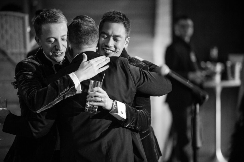 A black and white photograph capturing an emotional embrace among friends at a 74 Wythe rooftop wedding. The image shows two of the wedding party members, laughing joyfully and hugging tightly, celebrating the special moment together. 
