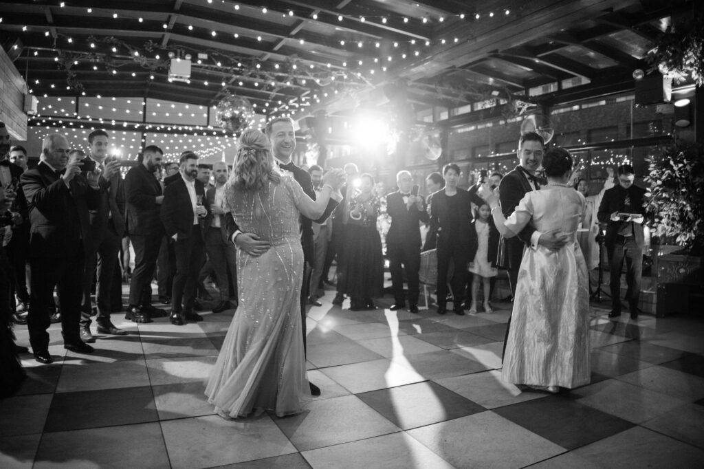 A touching black and white photograph from a 74 Wythe rooftop wedding capturing a special moment where the grooms share a dance with their mothers. Surrounded by guests and under a canopy of twinkling lights, the pairs sway to the music, creating a heartfelt and memorable scene. 