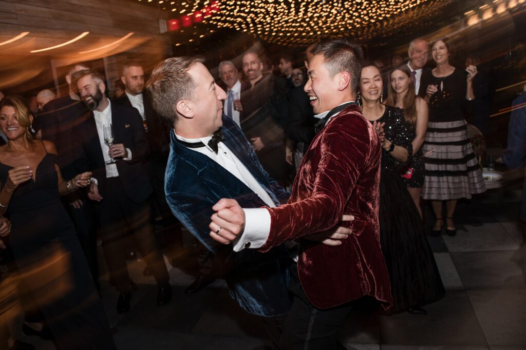 A lively photograph from a 74 Wythe rooftop wedding, capturing a joyful dance moment. The couple, dressed in stylish velvet tuxedos, laugh and dance together amidst a festive crowd of guests under twinkling lights, creating a vibrant and celebratory atmosphere. 