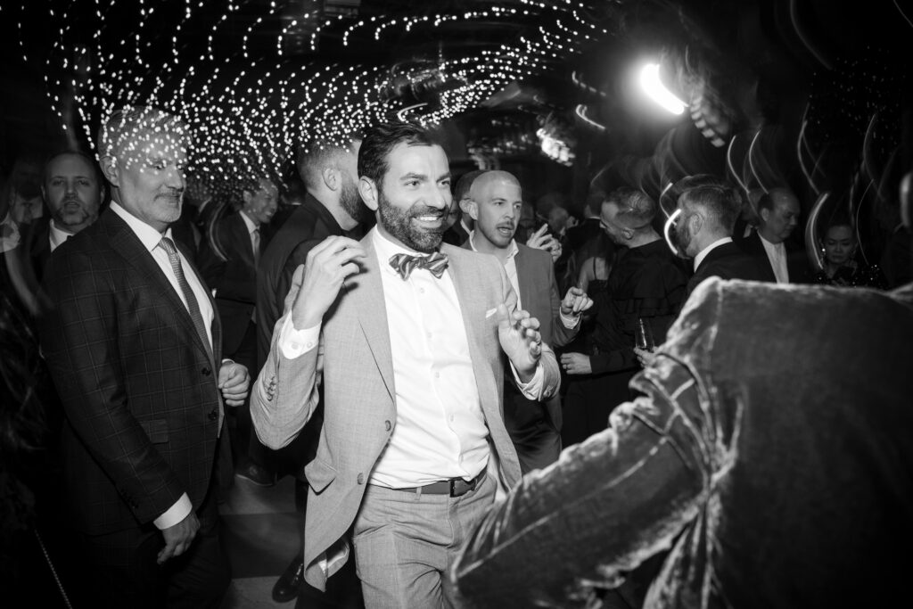 A black and white photograph capturing a lively dance moment at a 74 Wythe rooftop wedding. In the foreground, a guest with a joyful expression dances energetically, dressed in a light gray suit and a playful bow tie. The sparkling lights overhead add a magical touch to the festive atmosphere, with other guests enjoying the celebration in the background. 