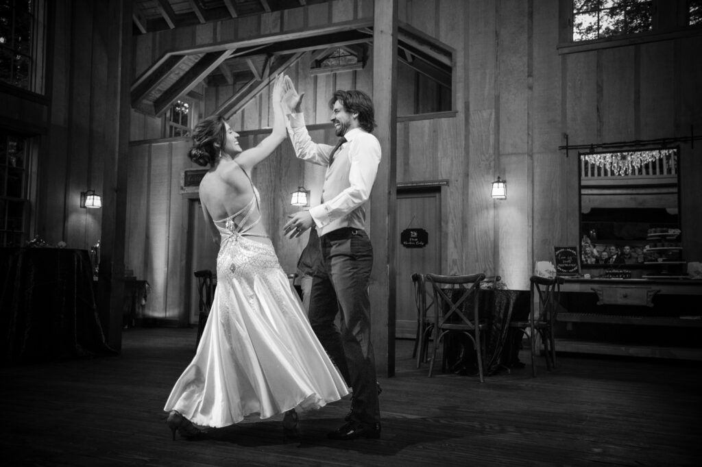 A couple dances together at a Nestldown wedding in a wooden venue, with the woman wearing a flowing dress and the man in a dress shirt and vest, as they engage in a high-five.