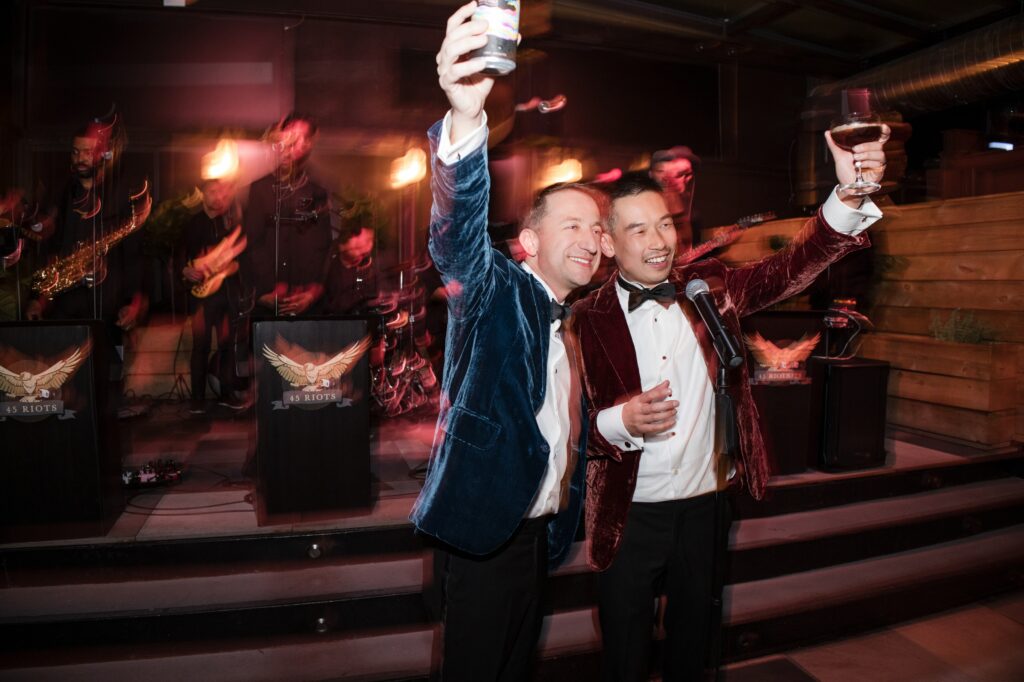 A vibrant photograph capturing a celebratory toast at a 74 Wythe rooftop wedding. The couple, dressed in elegant velvet tuxedos, joyfully raises their glasses in a toast, while one of them holds up a phone, possibly capturing the moment. The lively band performs in the background, enhancing the festive atmosphere of the celebration. 