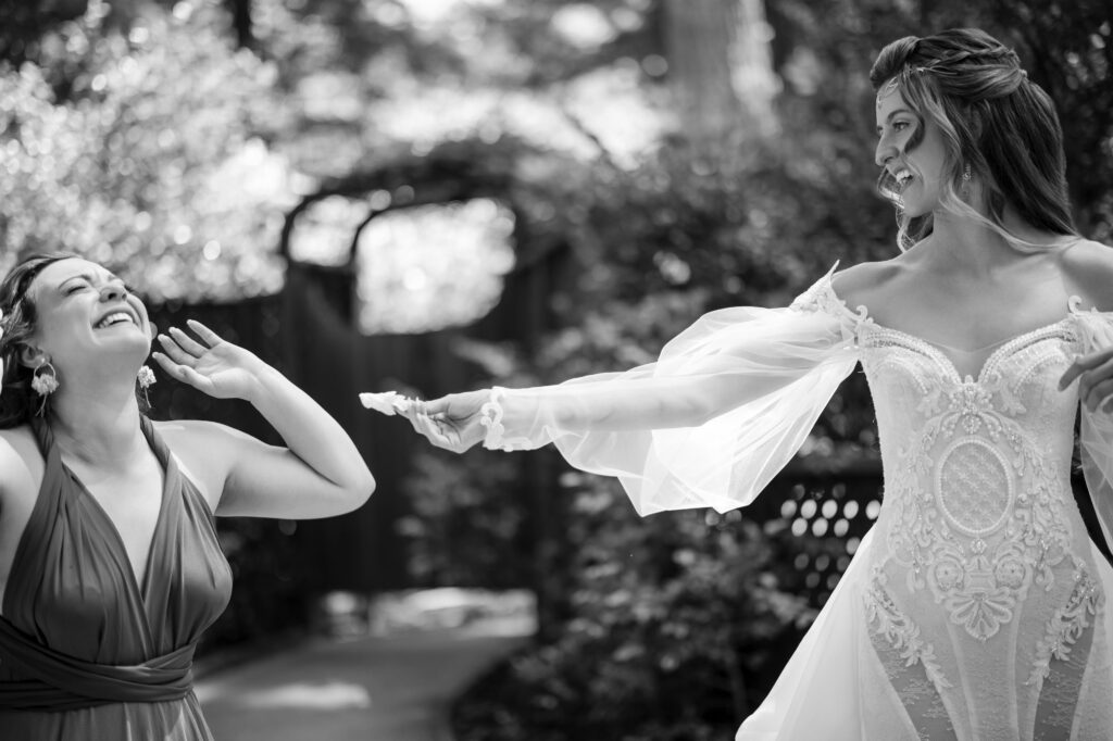 A bride in an ornate gown reaches out playfully to a woman in a dress, both smiling amid the enchanting landscape of a Nestldown wedding.