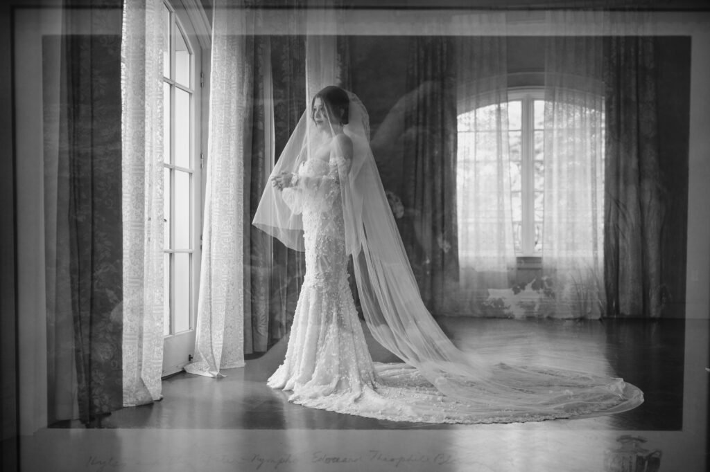 A bride in a wedding gown and veil stands by a large window at Park Chateau, holding a bouquet. The room features long curtains with sunlight filtering in. The image has a double-exposure effect, adding a layered look.
