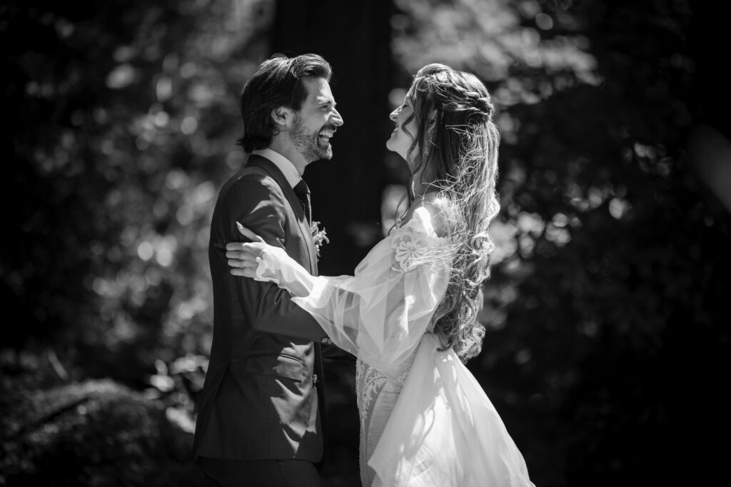 Black and white photo of a couple in wedding attire standing outdoors at Nestldown, smiling and looking at each other while holding hands.