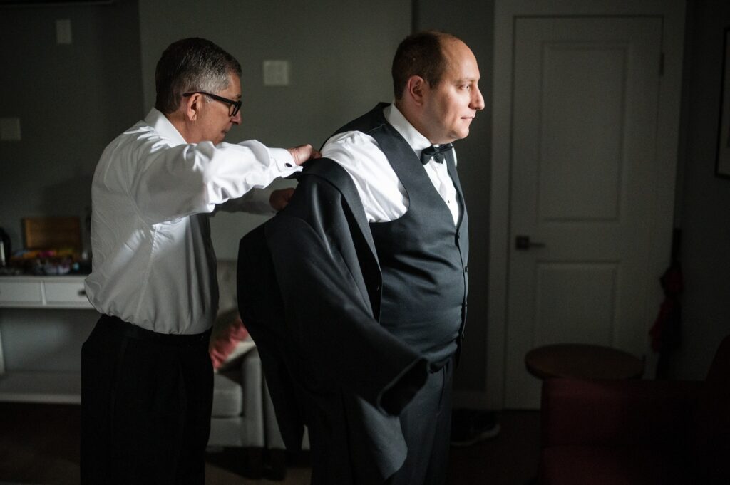 groom gets ready for wedding as another person helps put on his jacket