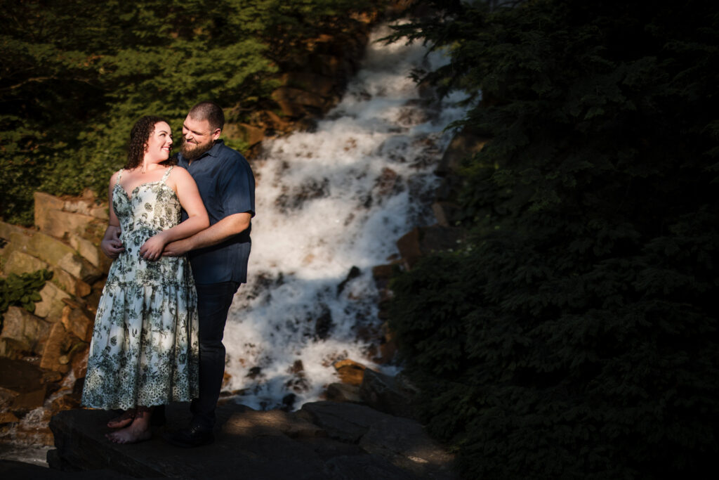 A couple stands closely together, smiling and embracing during their Longwood Gardens engagement session, with a waterfall and greenery in the background.