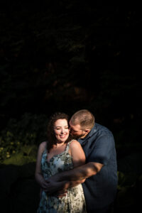 A man and woman stand close in a dimly lit natural setting at Longwood Gardens, with the man gently kissing the woman's cheek. The woman smiles and holds his arms wrapped around her in this intimate engagement session.