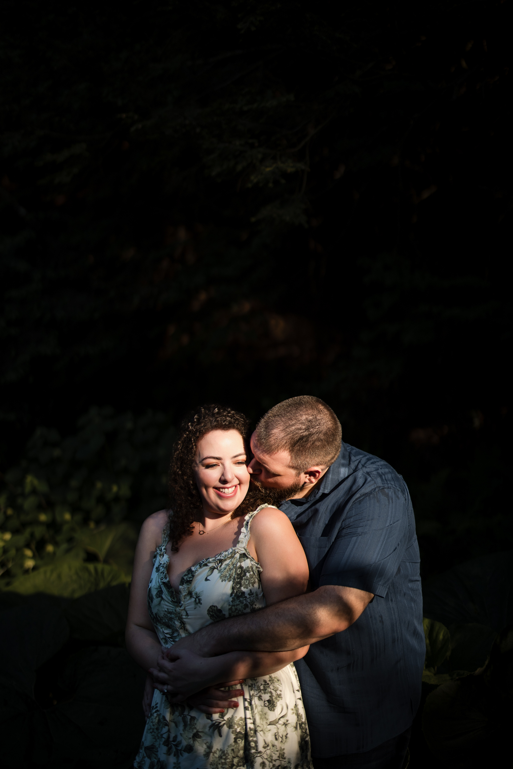 A man and woman stand close in a dimly lit natural setting at Longwood Gardens, with the man gently kissing the woman's cheek. The woman smiles and holds his arms wrapped around her in this intimate engagement session.