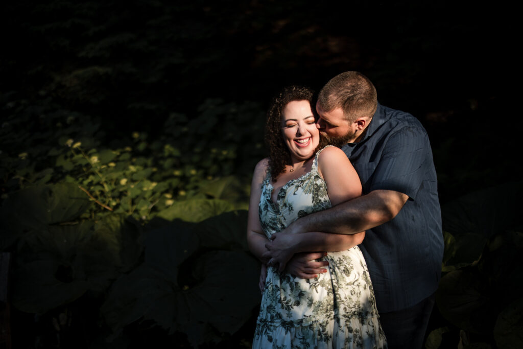 A man embraces a smiling woman from behind in a garden, both illuminated by soft light during their Longwood Gardens engagement session.