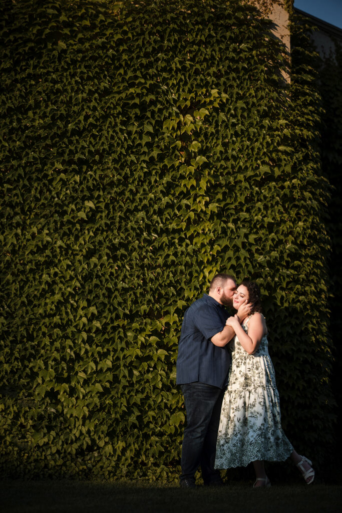 A couple stands close, embracing in front of a large, ivy-covered wall at Longwood Gardens. The man kisses the woman's forehead as she smiles, creating a perfect moment for their engagement session.