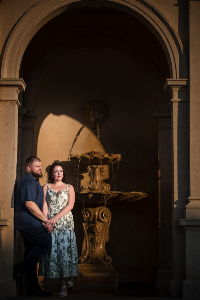 A couple stands together in front of a stone fountain under an archway at Longwood Gardens, with soft sunlight illuminating the scene. The woman wears a floral dress, and the man is in a navy shirt. This serene moment captures the essence of their engagement session beautifully.