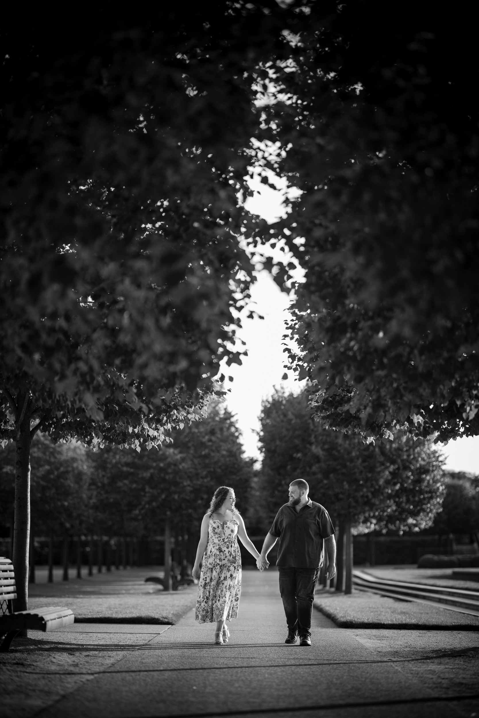A couple holding hands walks down a tree-lined pathway in a park during their Longwood Gardens engagement session. The image is in black and white.