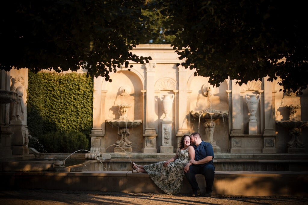 A couple sits closely together on the edge of a fountain in a historic stone courtyard, with ornate sculptures and lush greenery in the background, capturing the romance of their Longwood Gardens engagement session.