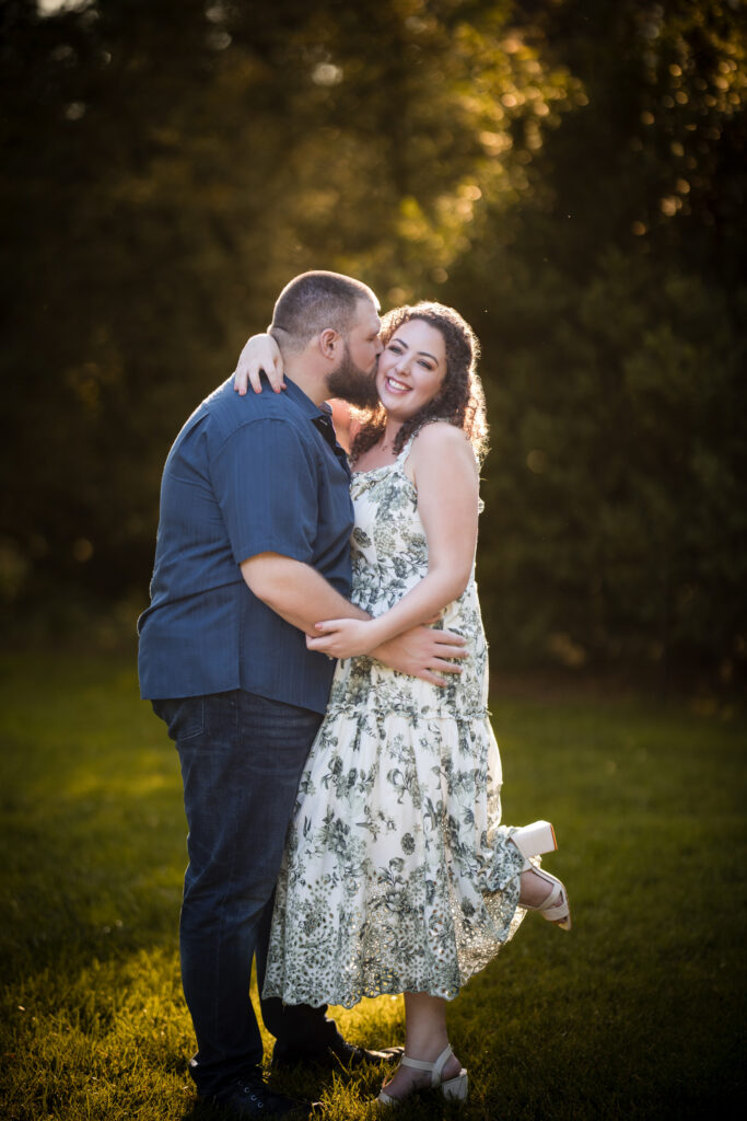 A man and woman stand outdoors at Longwood Gardens, embracing and smiling. The man is wearing a blue shirt and jeans, while the woman dons a white floral dress. Sunlight filters through the trees in the background, capturing the magic of their engagement session.