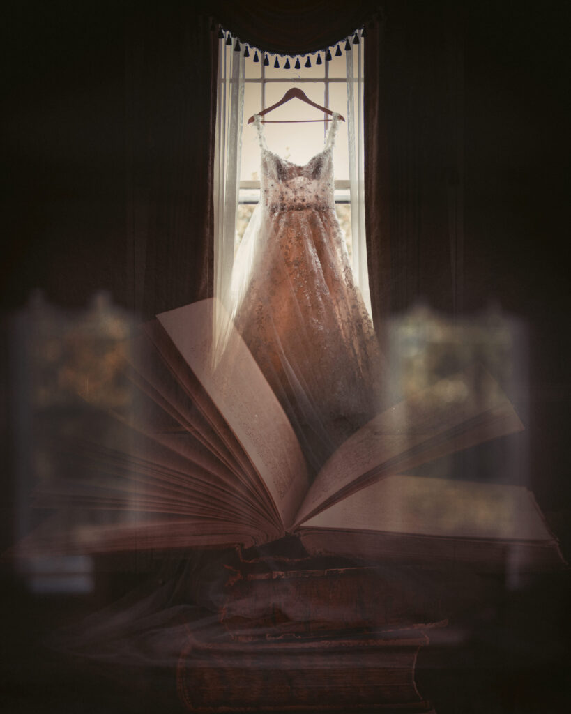 A wedding dress hangs on a hanger in front of a window. An open book is positioned in the foreground, partially blurred.