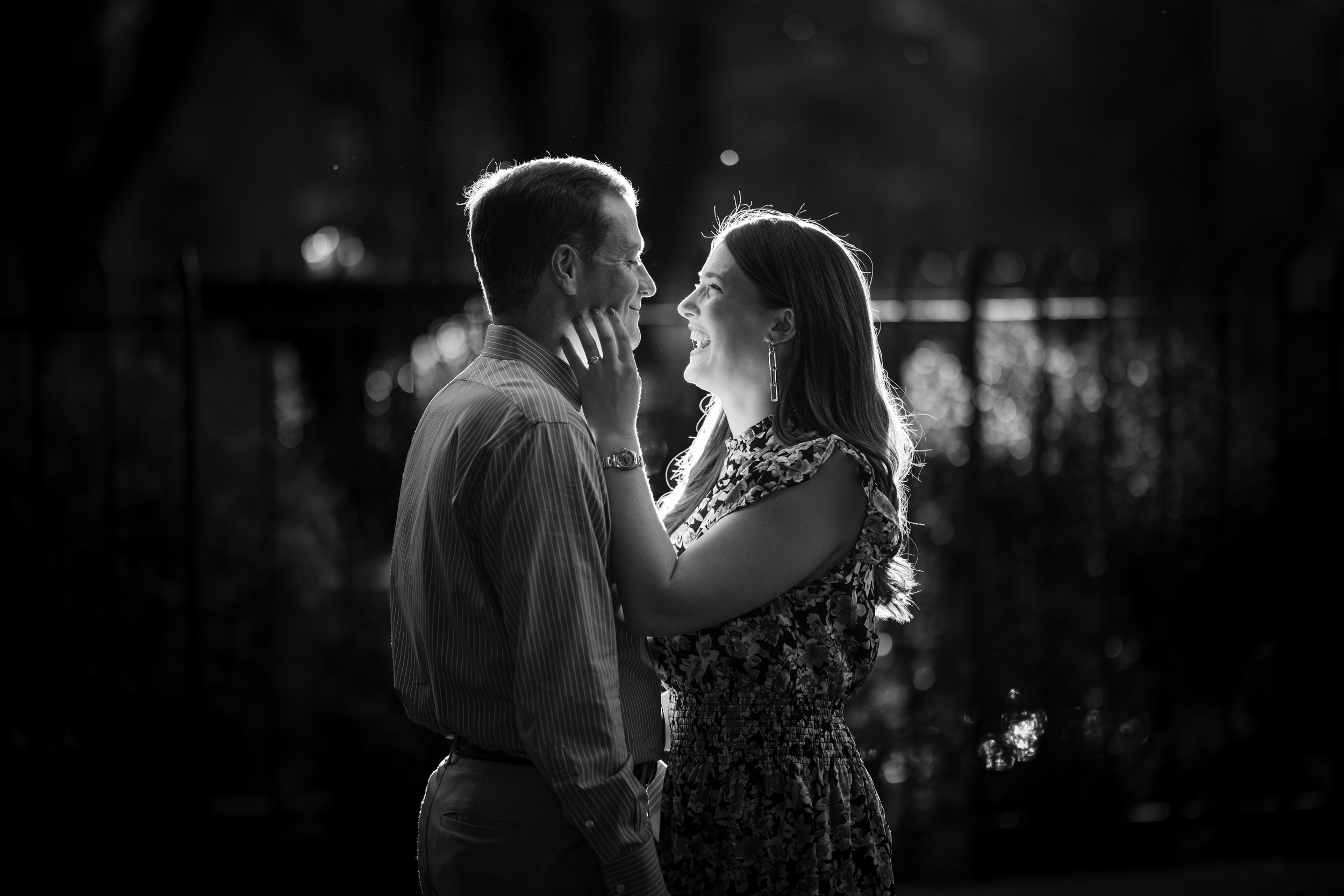 A man and a woman stand facing each other outdoors, smiling and touching each other's faces, with sunlight shining from behind, showcasing the perfect moment captured after choosing a location for your engagement photos.