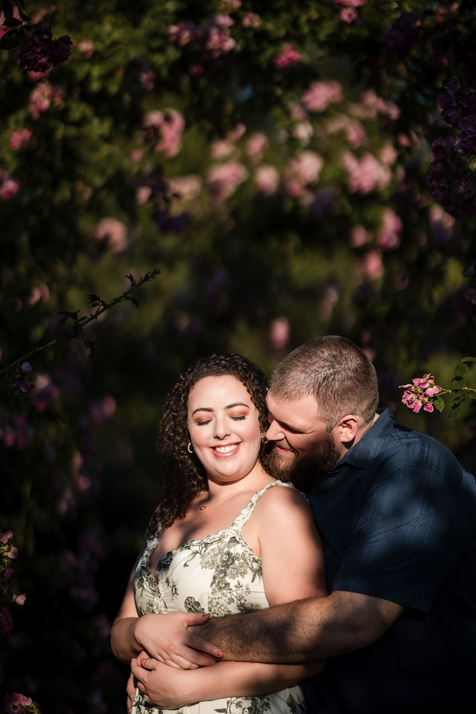 A couple stands closely, smiling with eyes closed, amidst a backdrop of blooming flowers and greenery during their Longwood Gardens engagement session.