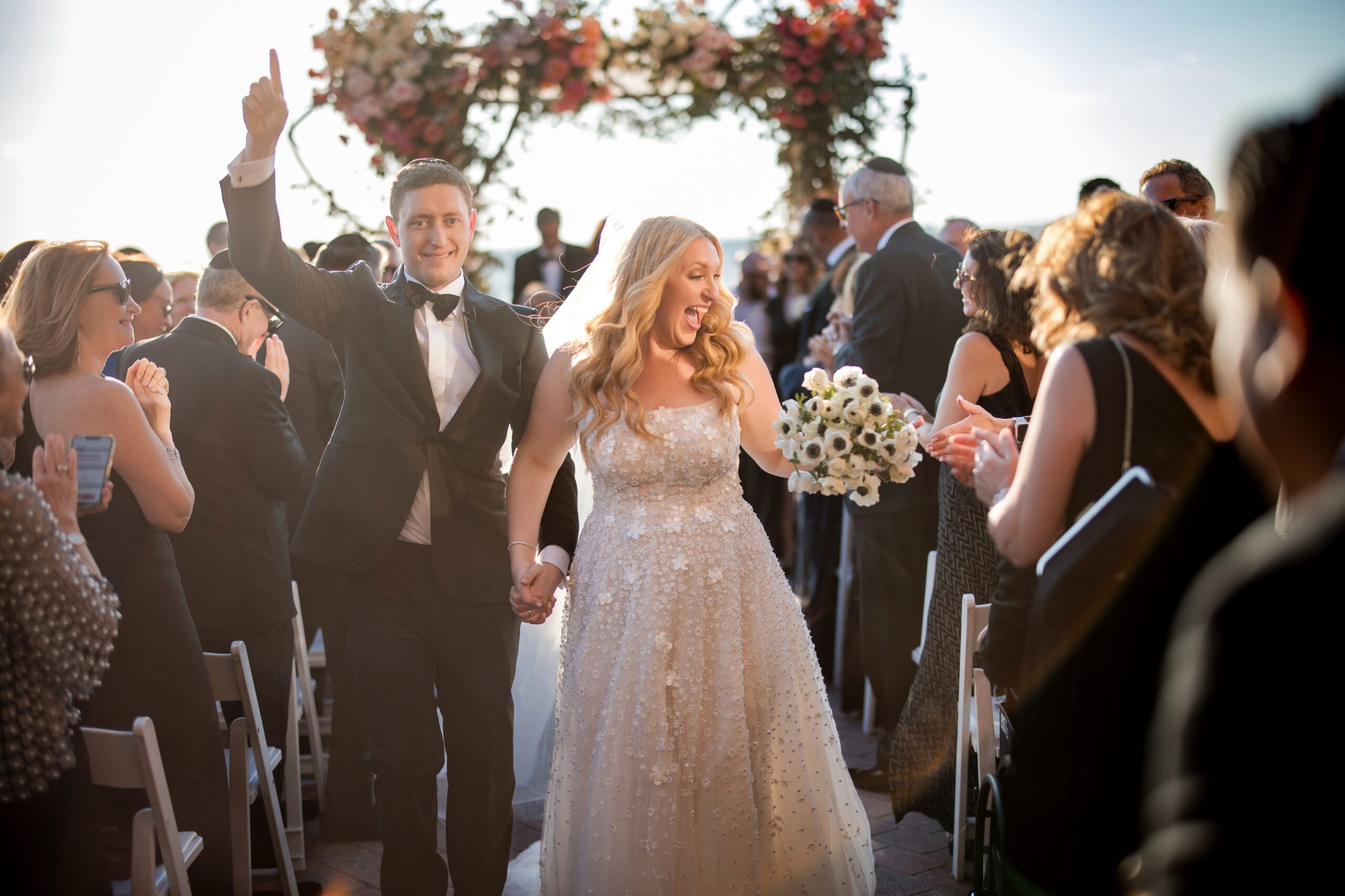 A bride and groom walk down the aisle after their Liberty Warehouse wedding, holding hands and smiling, surrounded by applauding guests. A floral archway is visible in the background.