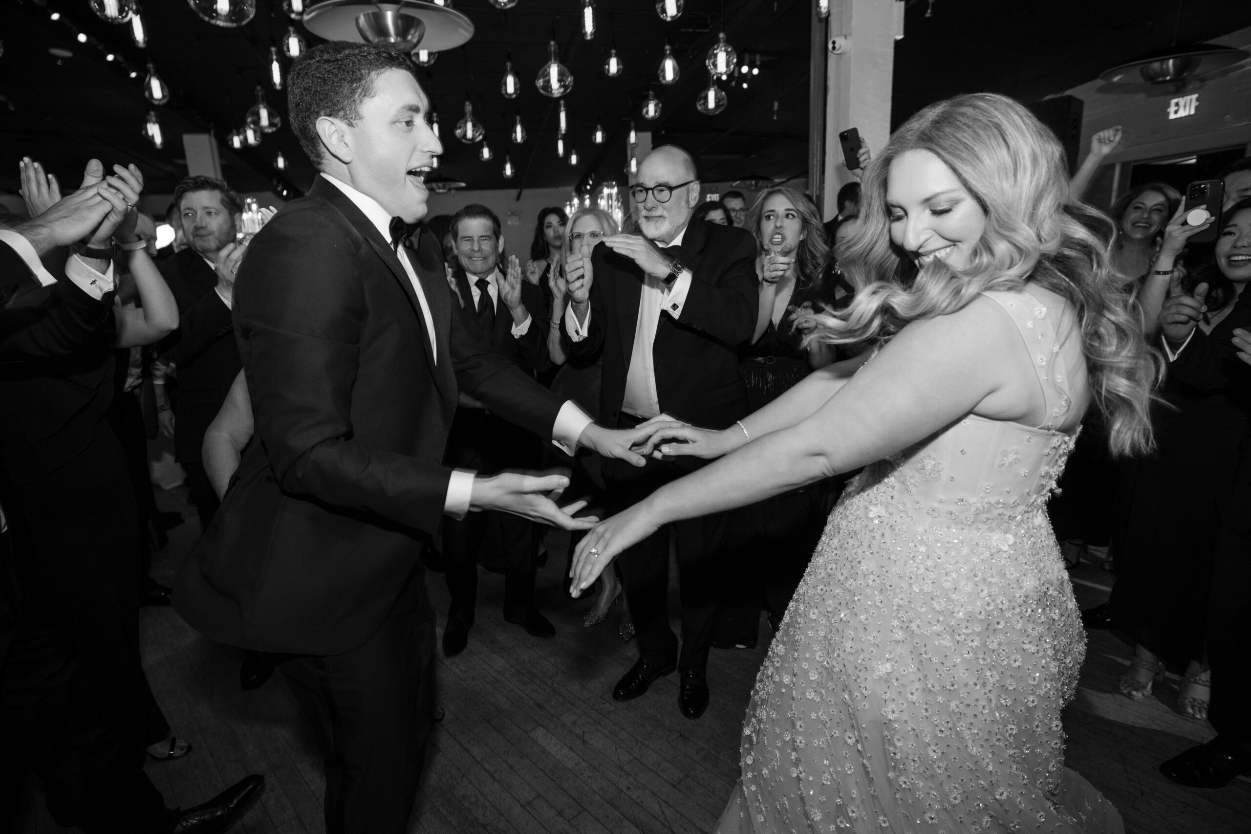 A bride and groom dance joyfully at their Liberty Warehouse wedding reception as guests surrounding them clap and cheer. The event takes place in a well-lit venue with hanging lights.