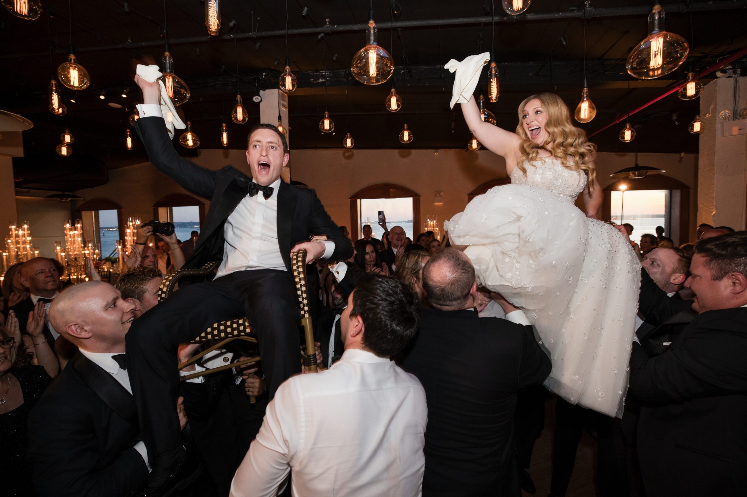 At a Liberty Warehouse wedding, a bride and groom are lifted in chairs during the celebration, surrounded by guests in formal attire. Hanging lights illuminate the festive scene.