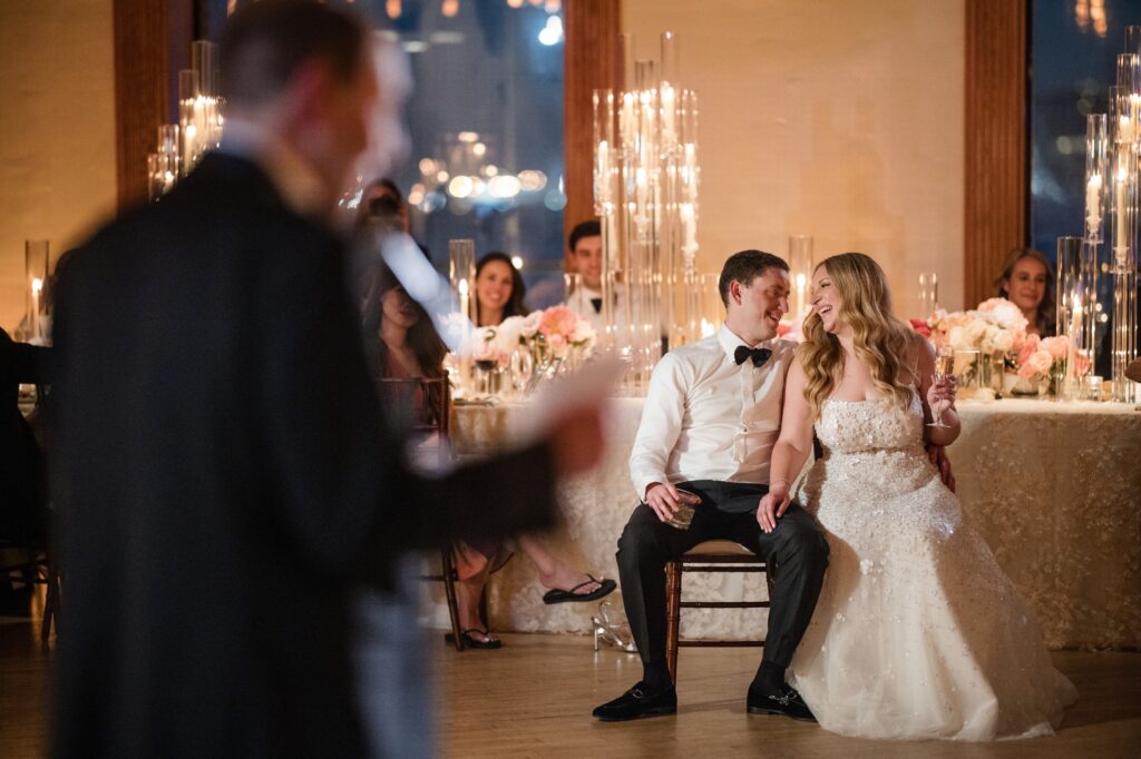 A couple, dressed in formal attire, sits close together and smiles at each other while listening to a speech at their Liberty Warehouse wedding. Guests and decorative floral arrangements are visible in the background.