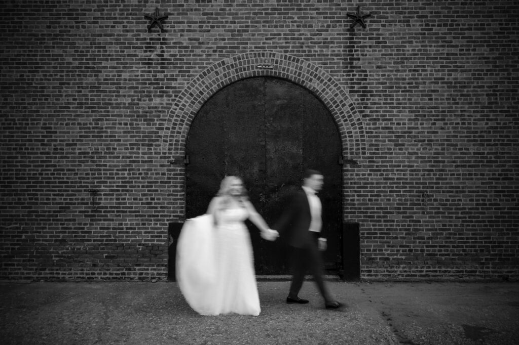 A bride and groom walk hand-in-hand in front of a large arched metal door set in a brick wall, reminiscent of a classic Liberty Warehouse wedding. The image is black and white, with both figures slightly blurred due to motion.