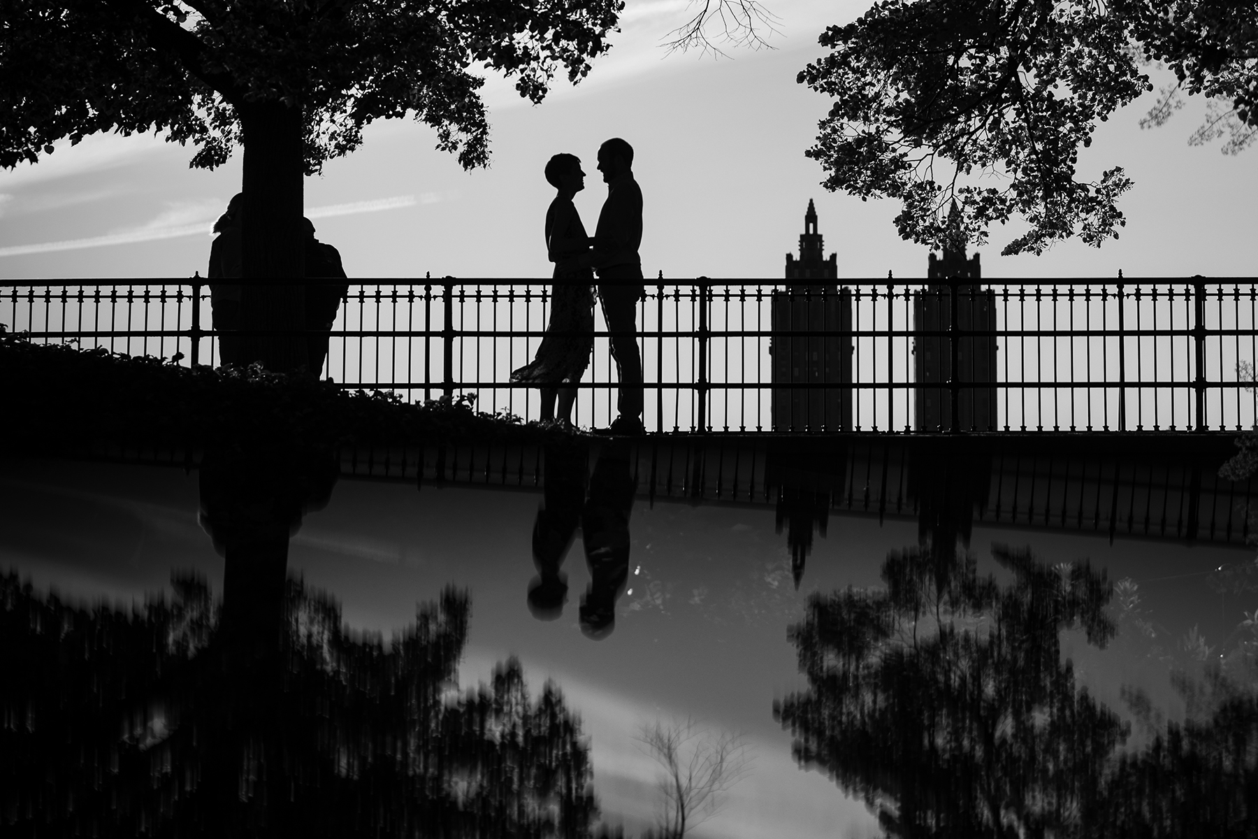 Choosing a location for your engagement photos, imagine a silhouetted couple standing together on a bridge with trees and buildings in the background, their reflection visible in the water below.