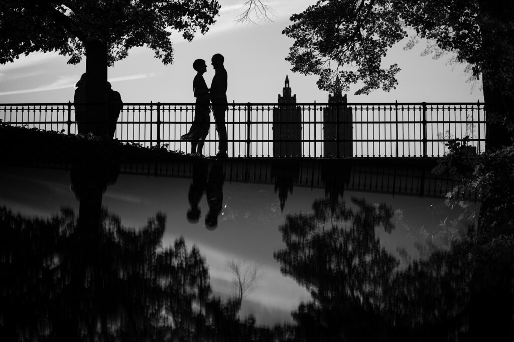 Silhouetted couple stands on a bridge, holding hands amidst trees. Their reflection is visible in the water below. Buildings and a clear sky are in the background—an exquisite example of choosing a location for your engagement photos.