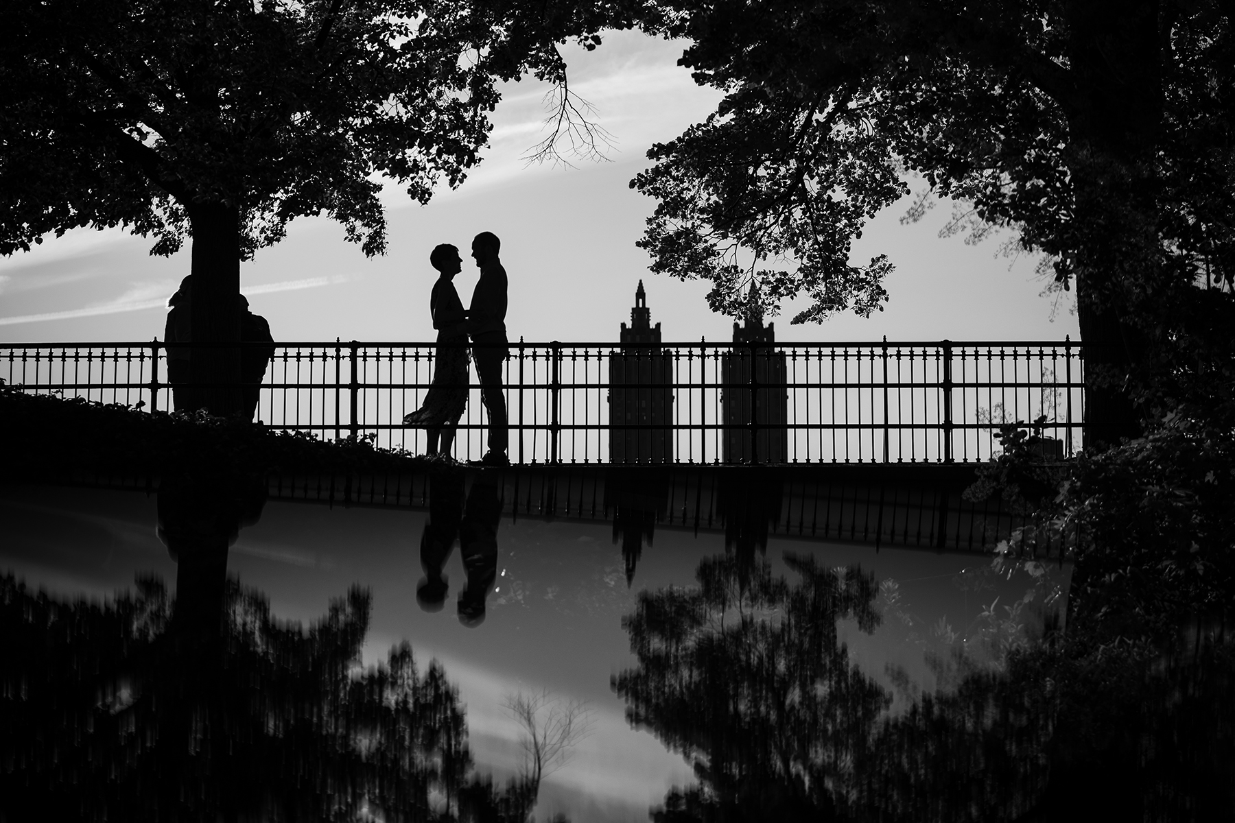 Silhouetted couple standing on a bridge surrounded by trees, with a city skyline in the background and their reflection visible in a body of water below—an idyllic scene perfect for choosing a location for your engagement photos.