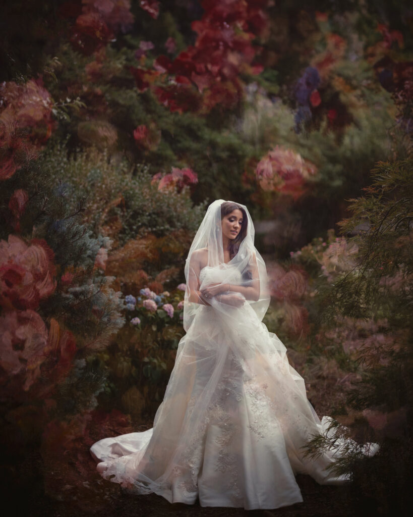 A bride in a white wedding dress and veil stands outdoors surrounded by vibrant flowers and foliage, perfectly capturing the beauty of a summer wedding.