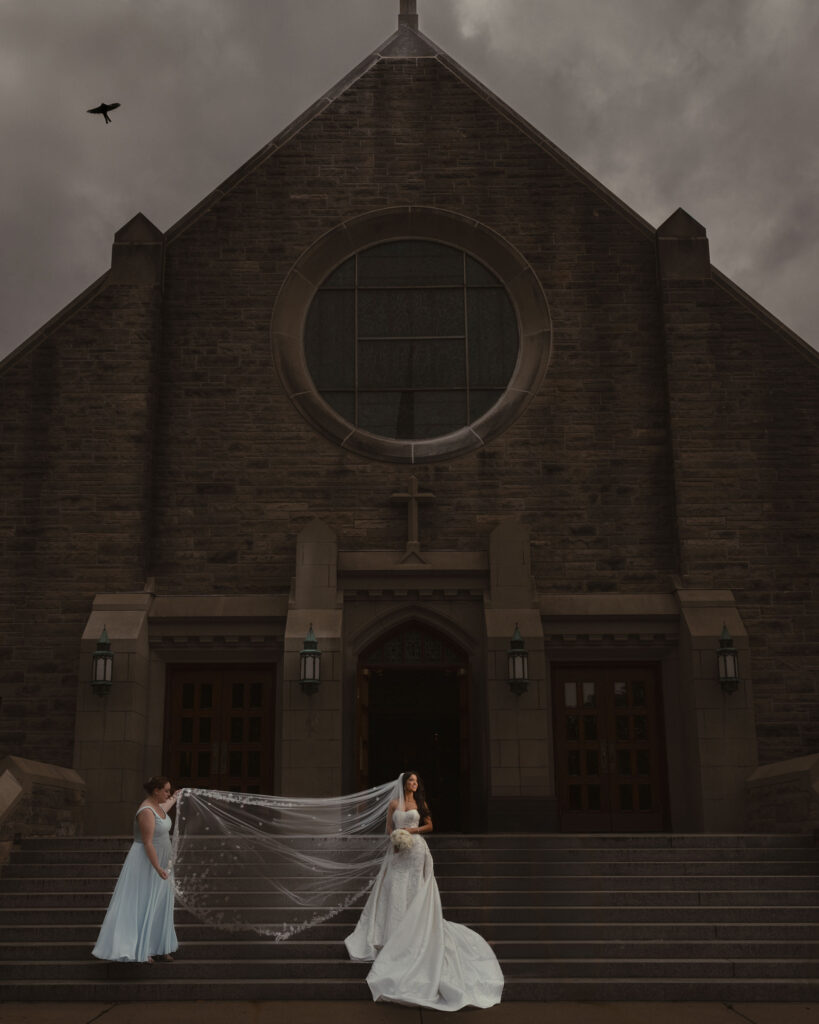 A bride in a white dress stands on the steps of a large stone church, accompanied by attendants adjusting her long veil. On this summer wedding day, a black bird is flying in the cloudy sky above. This bride used Tiktok to find her wedding photographer.