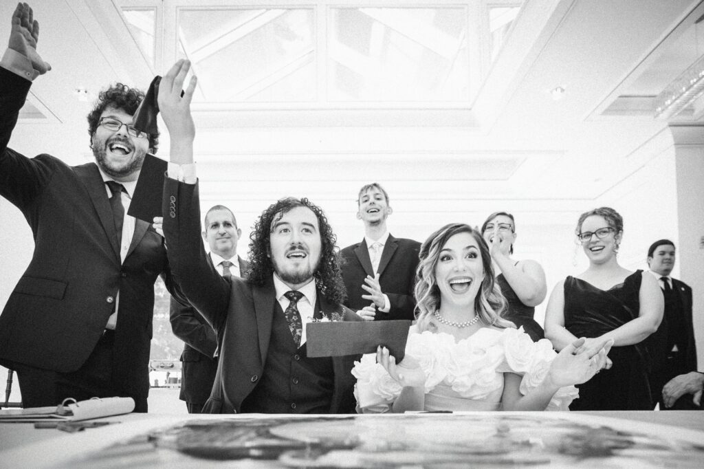 A group of people in formal attire celebrate indoors, with one man and woman seated at a table in the center, raising their hands in excitement, capturing the joy of a luxurious summer NJ wedding.