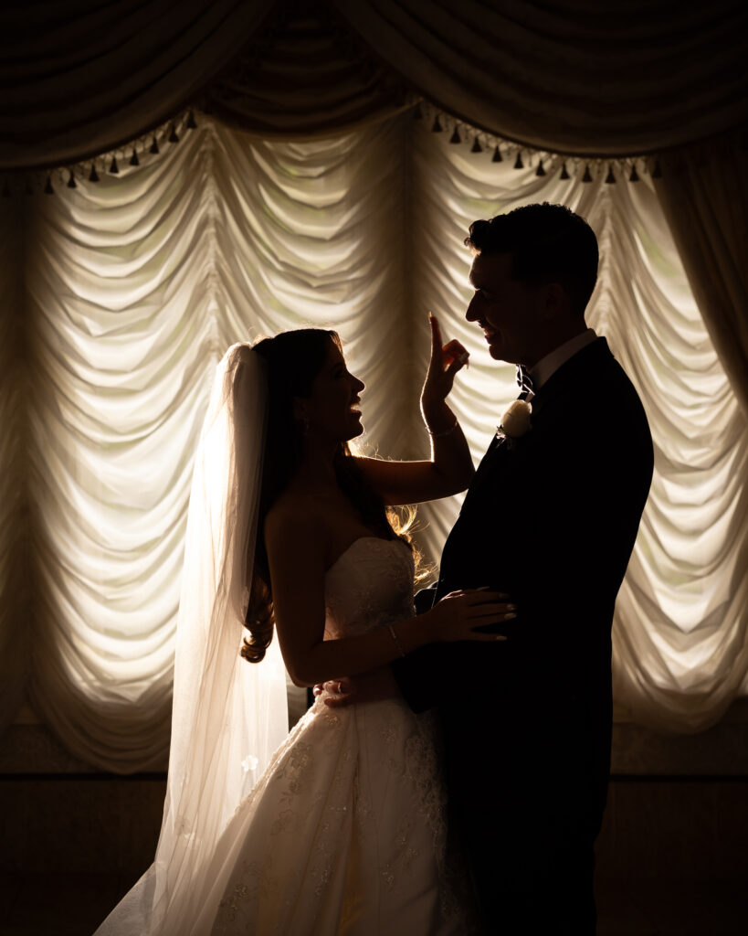 Silhouetted bride and groom share a moment in front of softly lit, draped curtains during their summer wedding. The bride is reaching up to the groom's face while the groom looks at her.