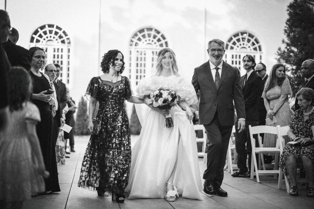 A bride in a white dress walks down the aisle with a man and woman on either side of her at a luxury summer NJ wedding, surrounded by guests in formal attire.