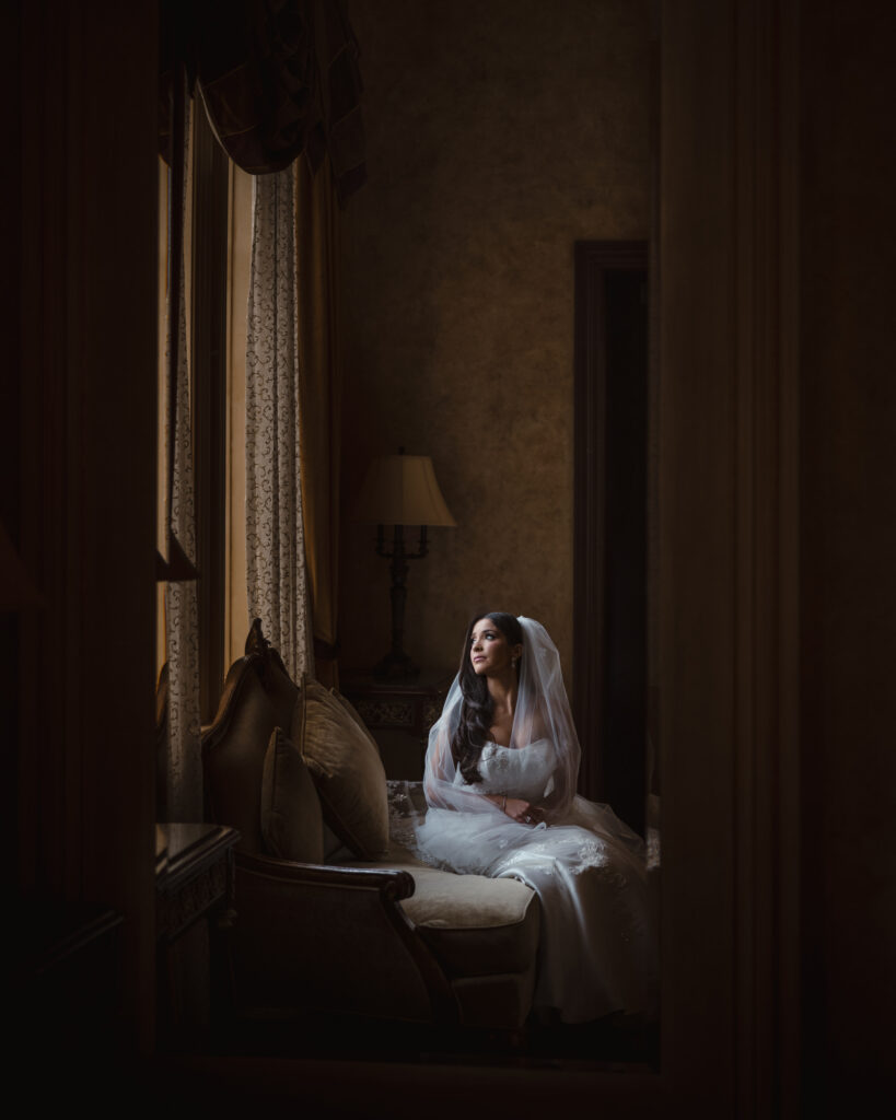 A woman in a white wedding dress and veil sits on a couch, gazing out of a window in the dimly lit room, lost in thoughts of her summer wedding.