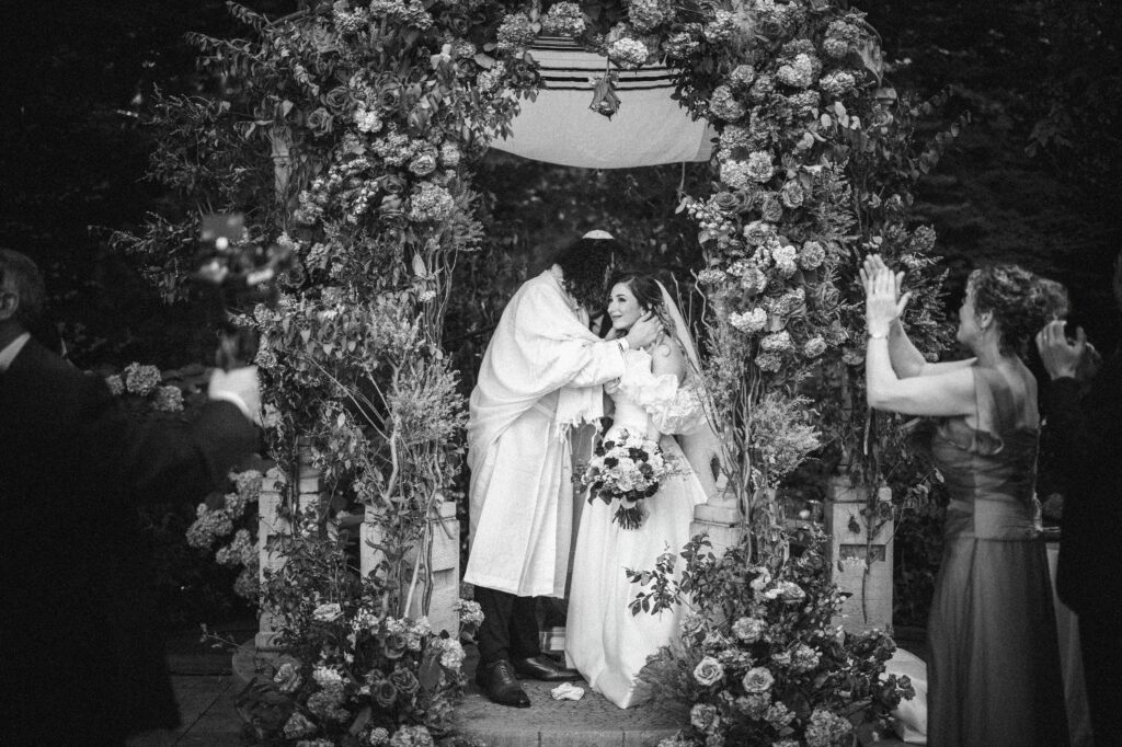 A bride and groom share an embrace under a floral arch during their luxury summer NJ wedding. Guests are clapping in the background.