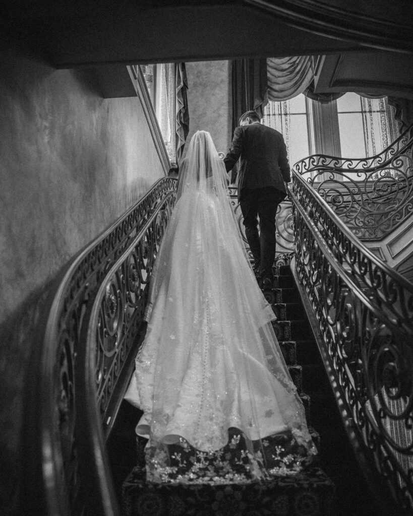 A bride and groom ascend a grand staircase. The bride wears a long, flowing gown and veil, perfect for their summer wedding, and the groom is dressed in a suit. The staircase has ornate railings and leads towards a large window.