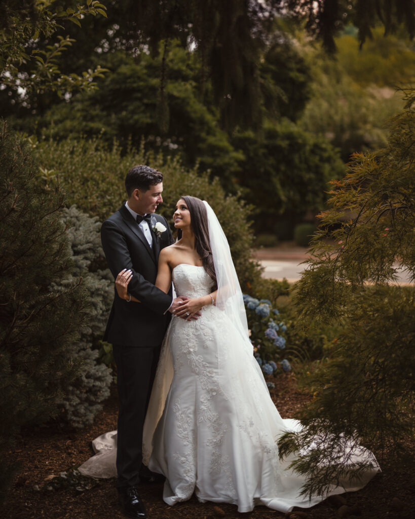 A bride in a white gown and a groom in a black tuxedo stand embracing in a lush garden setting, surrounded by greenery, capturing the essence of a perfect summer wedding.