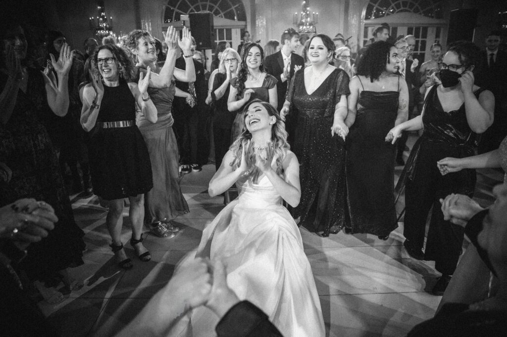 At a luxury summer NJ wedding, a bride in a white dress dances joyfully on the floor, surrounded by guests in formal attire, all clapping and smiling.