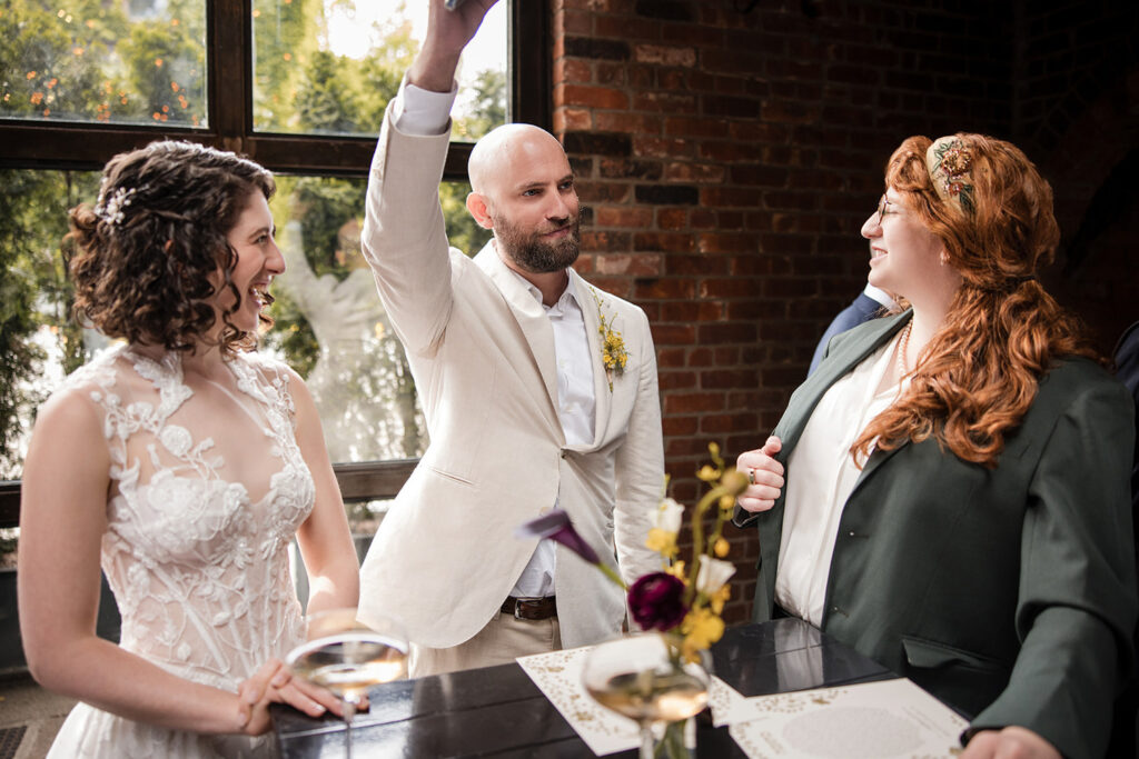 The groom raises his hand triumphantly while standing next to the smiling bride, who is holding a glass of wine, and a woman with red hair and glasses dressed in a green jacket. The scene is set indoors with brick walls and a large window offering a view of greenery outside. The atmosphere appears celebratory and joyful.