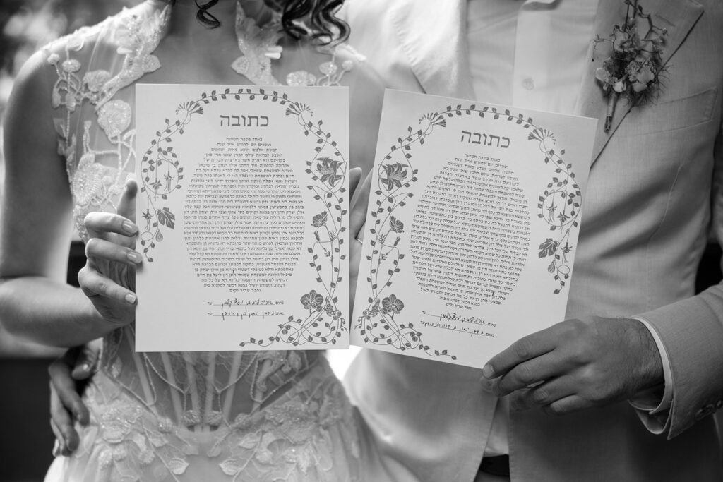 A bride and groom hold up their ketubah, a traditional Jewish marriage contract, decorated with floral illustrations. The bride's intricate lace dress and the groom's light suit jacket are partially visible as they display the document, which is written in Hebrew. The image captures a significant and formal moment in their wedding ceremony.