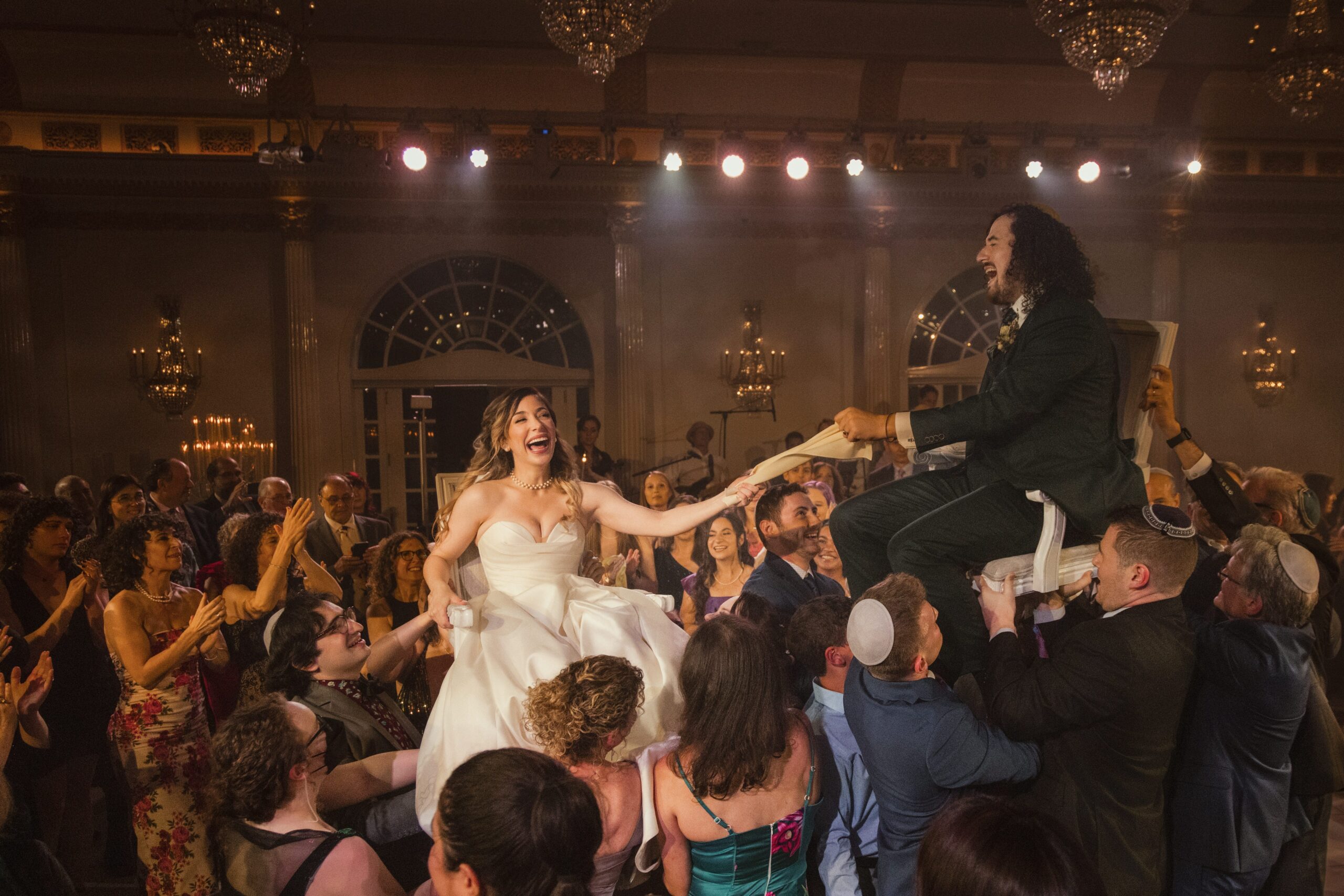 A bride and groom are lifted on chairs during a luxurious summer wedding celebration in NJ, surrounded by cheering guests in a grand ballroom.
