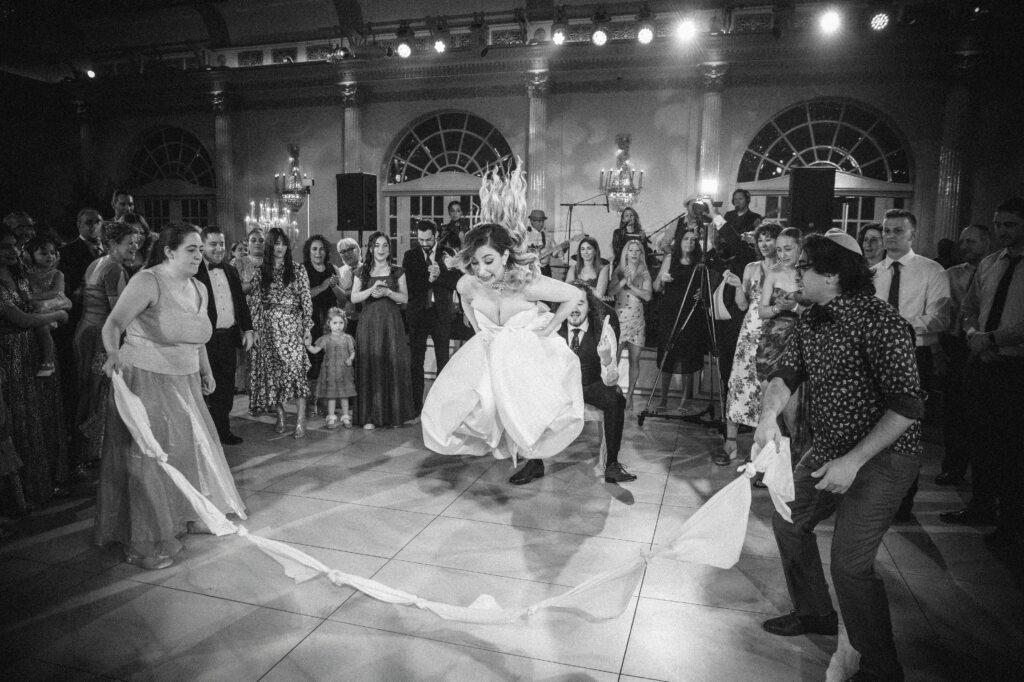 A bride jumps over a rope held by two people, surrounded by guests in a well-lit room with large windows. The luxury summer NJ wedding is festive and celebratory, as guests watch closely, some taking photos.