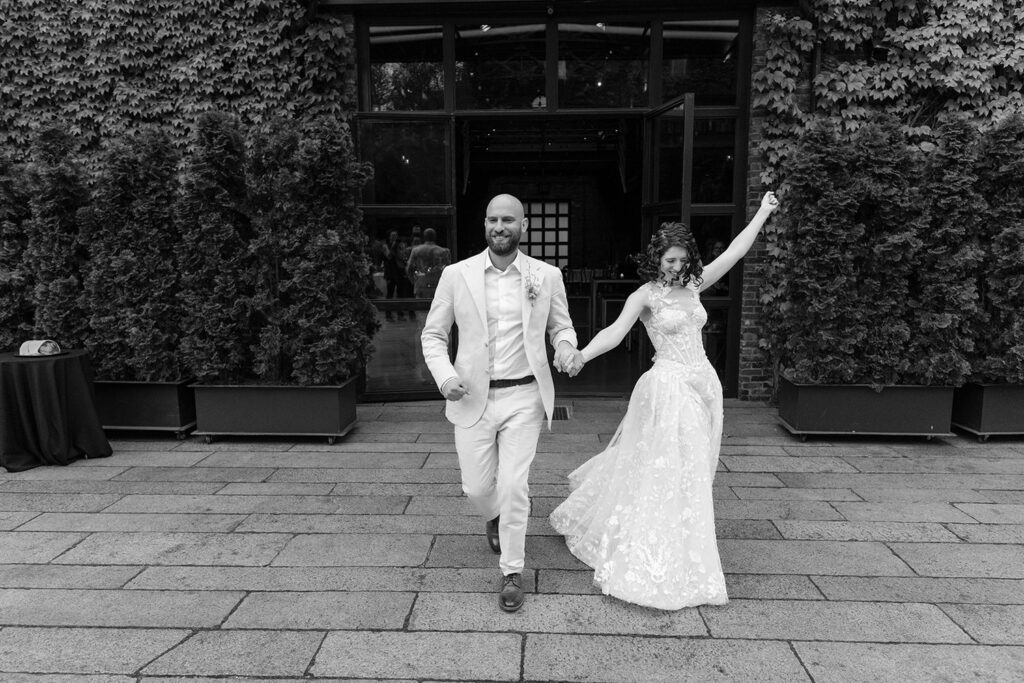 The bride and groom joyfully walk hand-in-hand, the bride raising her arm in celebration. The groom is in a light suit, and the bride wears a lace gown. They are exiting a building covered in ivy, with guests visible through the open doors behind them. The scene captures a moment of exuberance and happiness.