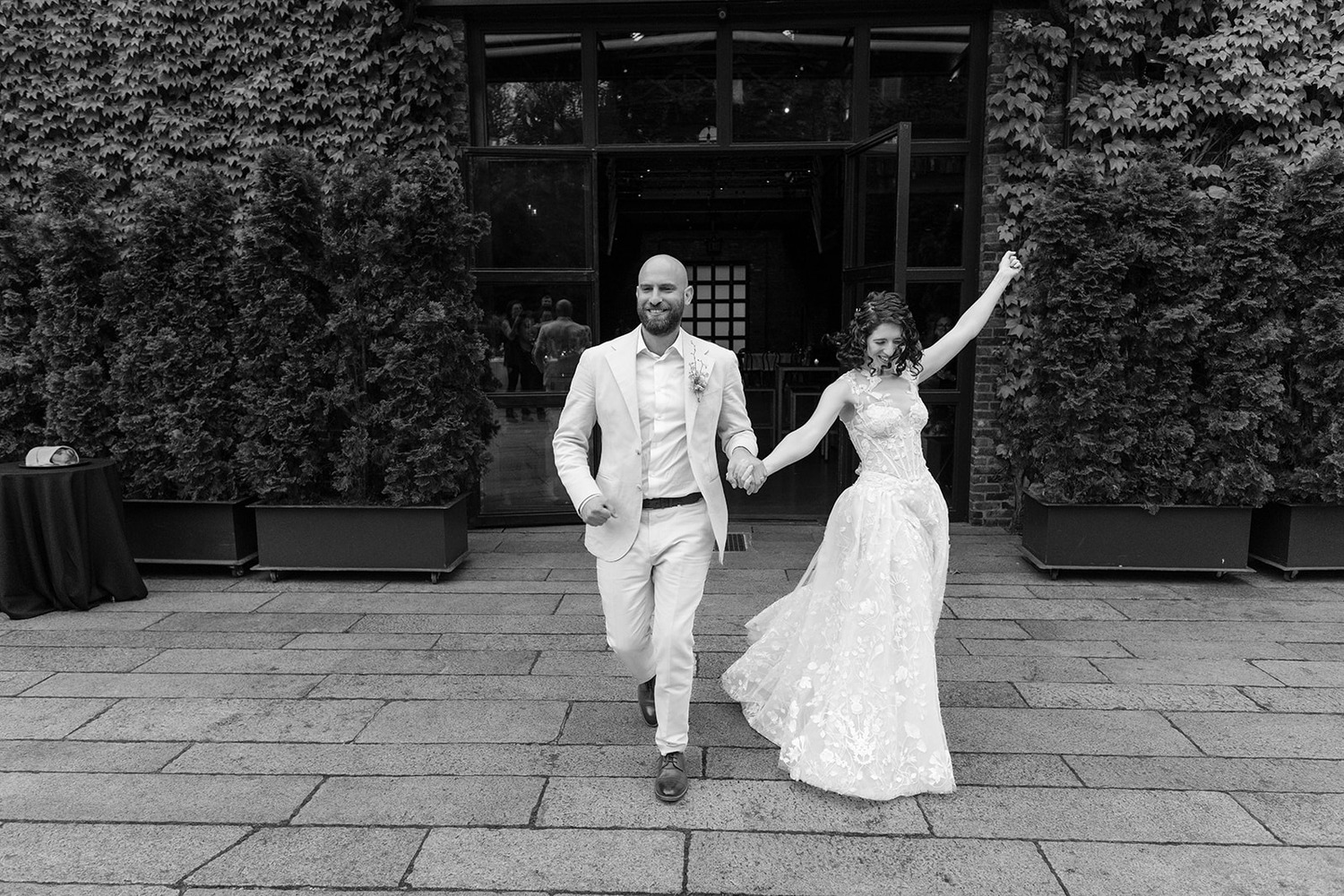 The bride and groom joyfully walk hand-in-hand, the bride raising her arm in celebration. The groom is in a light suit, and the bride wears a lace gown. They are exiting a building covered in ivy, with guests visible through the open doors behind them. The scene captures a moment of exuberance and happiness.