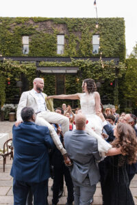 The bride and groom are lifted on chairs by their guests during the Hora dance, a traditional Jewish wedding celebration. The groom, in a light suit, and the bride, in a lace gown, reach out to hold hands as they are elevated above the crowd. The backdrop features an ivy-covered building and festive string lights, with smiling guests surrounding them in celebration.