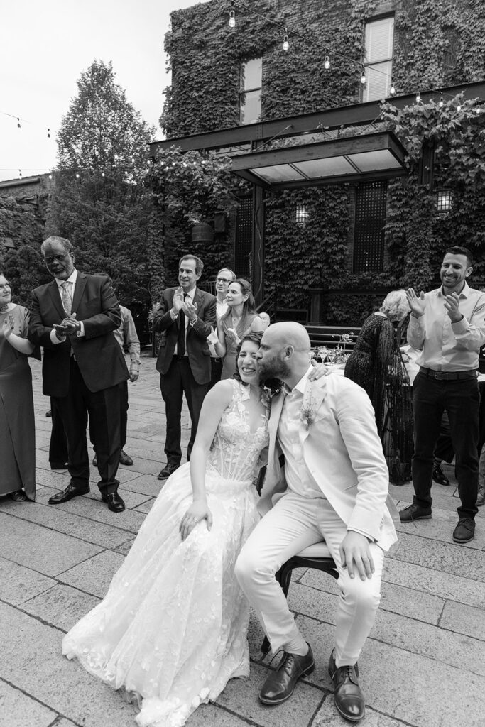 The bride and groom sit closely together, sharing a joyous moment while surrounded by applauding guests. The groom, in a light suit, kisses the bride, who is wearing a lace gown. The scene is set outdoors with ivy-covered walls and string lights, capturing a heartfelt celebration of their wedding day.