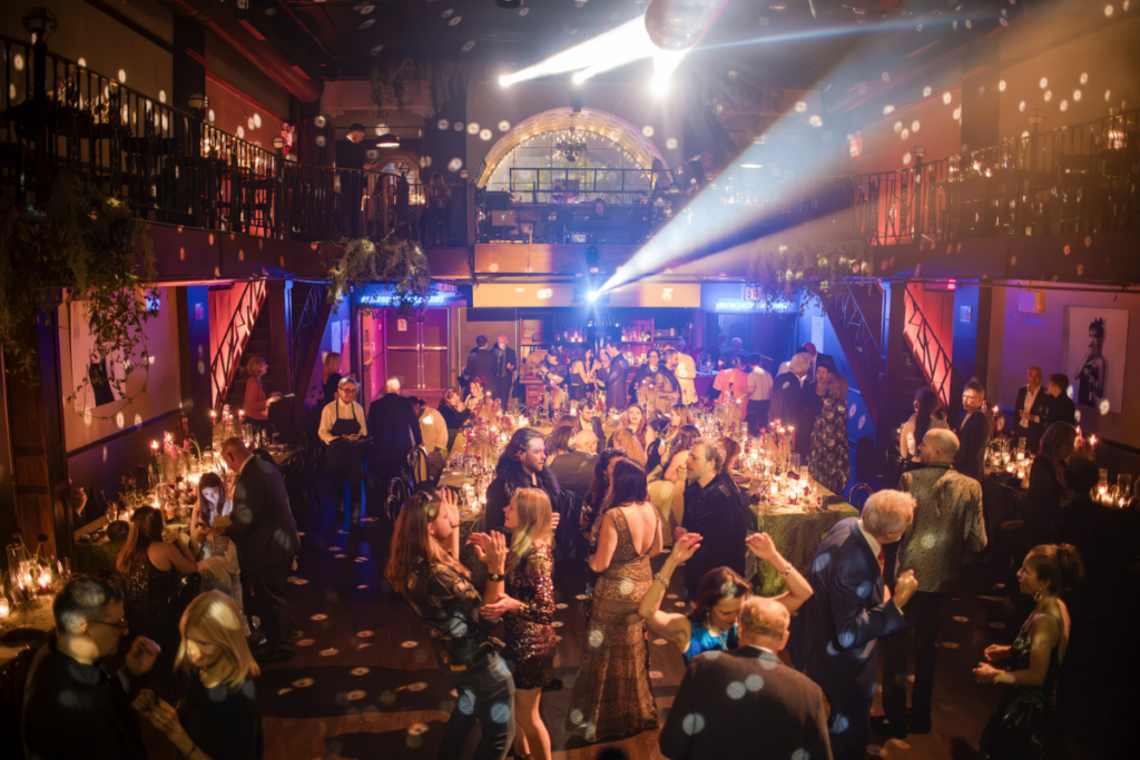 A large crowd of people in formal attire dance and socialize in a dimly lit ballroom, with tables, candles, and spotlights creating a festive atmosphere, perfect for kicking off an elegant Wedding Weekend.