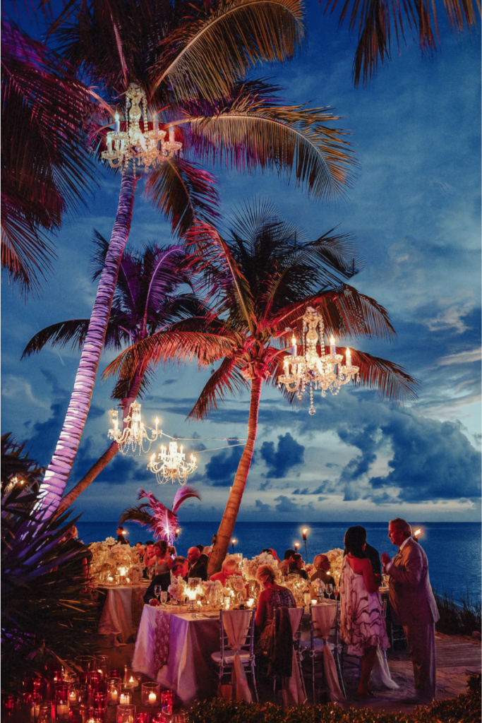 Wedding guests dining under palm trees adorned with chandeliers by a beach 